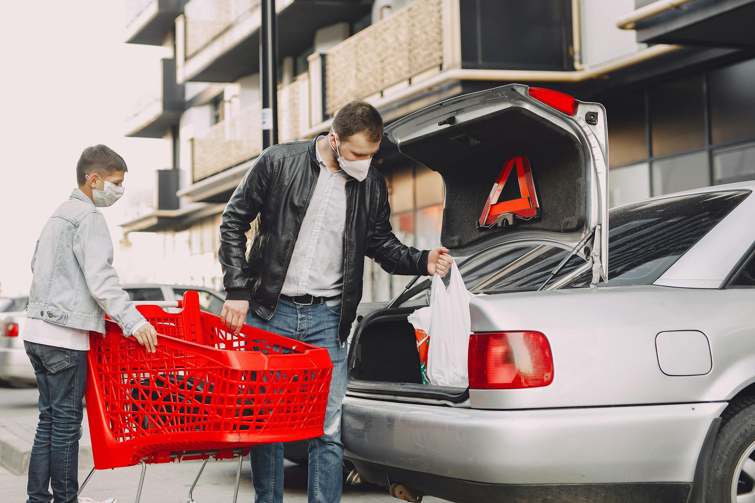 Man and boy in protective masks putting shopping bag in trunk